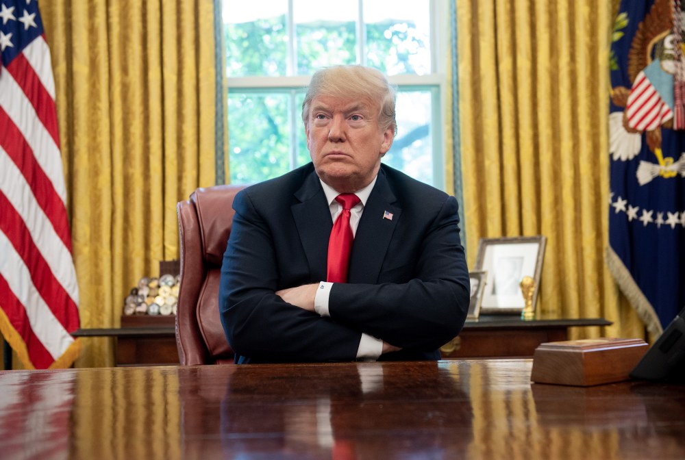 A photograph shows President Donald Trump sitting at his desk in the Oval Office with his arms crossed